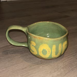May include: A green ceramic soup mug with a large handle. The word "SOUP" is painted in yellow on the side. The mug has a rustic, handcrafted appearance. The mug is likely made of stoneware.