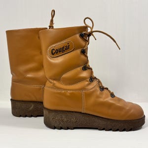 Puede incluir: Un par de botas de media caña color canela con acabado liso, cordones frontales con ojales metálicos y una suela gruesa de goma texturizada en color marrón oscuro. La palabra Cougar aparece impresa en un parche lateral y grabada en la suela.