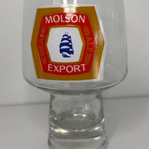May include: Clear glass beer glass with a wide top and a short stem. The glass features a Molson Export logo with a blue ship graphic and the words "Molson Export" in red and white.