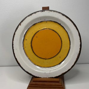 Può includere: Un piatto rotondo in ceramica con bordo bianco e un design concentrico di cerchi gialli e arancioni. Il piatto ha un bordo marrone scuro ed è esposto su un supporto di legno. Un pezzo di stoviglie vintage.