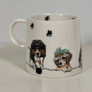 Puede incluir: Una taza de cerámica blanca con ilustraciones de dos perros beagle. Un beagle lleva un casco de hockey negro con la palabra HOCKEY impresa, mientras que el otro lleva gafas de montura blanca y una cinta para el pelo azul claro con lunares. Hay aves.