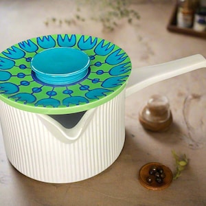 May include: White saucepan with a long handle and a decorative lid. The lid features a green and blue floral pattern with a turquoise center knob. The saucepan has vertical ridges and a pouring spout. A stylish kitchenware item.