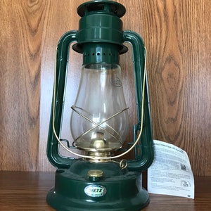 Könnte beinhalten: Eine grüne Kerosinlampe der Marke Dietz mit einer Glaskugel und einem Messingbrenner. Die Lampe steht auf einer Holzoberfläche mit Gebrauchsanweisung.