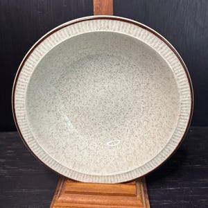Può includere: Una ciotola rotonda in ceramica maculata con interno grigio chiaro e bordo marrone. Il bordo presenta un design strutturato e a coste. La ciotola è esposta su un supporto in legno, mettendo in risalto la sua estetica vintage. Questa ciotola è adatta per servire cibo.