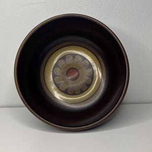 May include: A brown ceramic bowl with a floral design in the centre. The bowl has a dark brown exterior and a lighter brown and tan floral pattern in the centre. The floral pattern has a red centre.