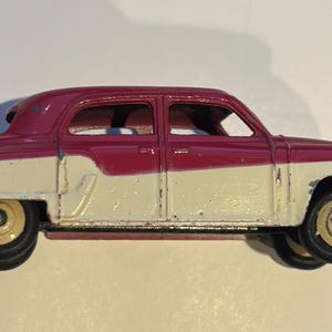 Könnte beinhalten: Ein Oldtimer-Spielzeugauto in einem zweifarbigen Farbschema aus Creme und Weinrot. Die klassische Limousine hat schwarze Räder mit goldfarbenen Zentren. Das Spielzeugauto hat ein detailliertes Design, einschließlich Fenster und Stoßstange. Ca. 15 cm lang.
