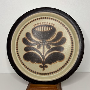 May include: A brown and cream ceramic plate with a stylized black floral design. The plate has a brown rim and a white background with a black floral design in the center.