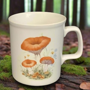 May include: White ceramic mug with a mushroom design. The mug features three orange mushrooms with white stems and brown leaves.