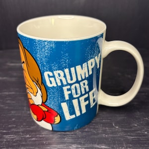 May include: White ceramic mug with a blue background and the words "GRUMPY FOR LIFE" in white. The mug features a cartoon character in brown, red, and white. The handle is white.
