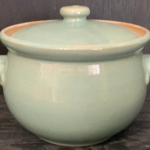 May include: A light green ceramic casserole dish with a lid and two handles. The dish has a slightly rounded shape and a smooth, glazed finish.