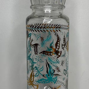 May include: A clear glass jar with a lid, decorated with a teal and gold floral pattern. The jar has a wide mouth and a rounded base.