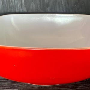 Pode incluir: Uma tigela quadrada de vidro vermelho e branco. A tigela é feita de Pyrex e tem um interior branco e um exterior vermelho.