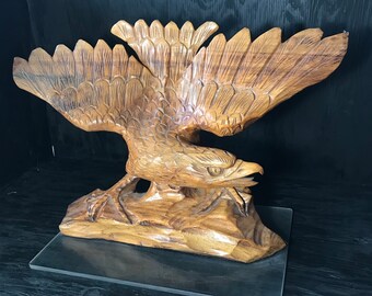 May include: A wooden eagle sculpture with detailed carving, perched on a base. The eagle's wings are spread wide, and its head is turned to the side. The sculpture is made of light brown wood.
