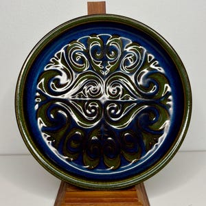 May include: A round ceramic plate with a blue centre and a green rim. The plate features a white and green ornate design in the centre. The plate is displayed on a wooden stand. This decorative plate is suitable for serving food.
