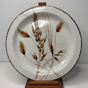 May include: A round, off-white ceramic plate with a brown rim and a wheat stalk design. The wheat stalks are in shades of brown and beige. The plate is displayed on a wooden stand.