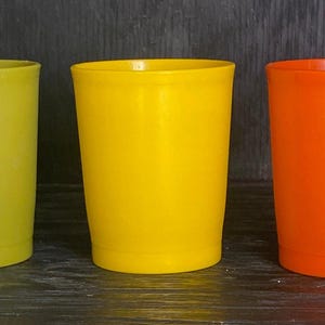 Puede incluir: Tres vasos de plástico de colores en fila. Los vasos son amarillo-verdosos, amarillos y naranjas. Los vasos son cilíndricos con un borde ligeramente ensanchado. Están sobre una superficie de madera oscura.