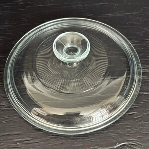 Puede incluir: Una tapa de vidrio transparente con un diseño circular y un pequeño asa redondeada. La tapa presenta una serie de crestas concéntricas que irradian desde el centro. Utensilios de cocina.