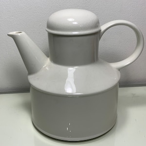 Może przedstawiać: Biały ceramiczny czajnik z zaokrąglonym korpusem i pokrywką. Czajnik ma dzióbek i uchwyt.