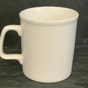 May include: A plain white ceramic mug with a handle. The mug is empty and sitting on a dark surface.