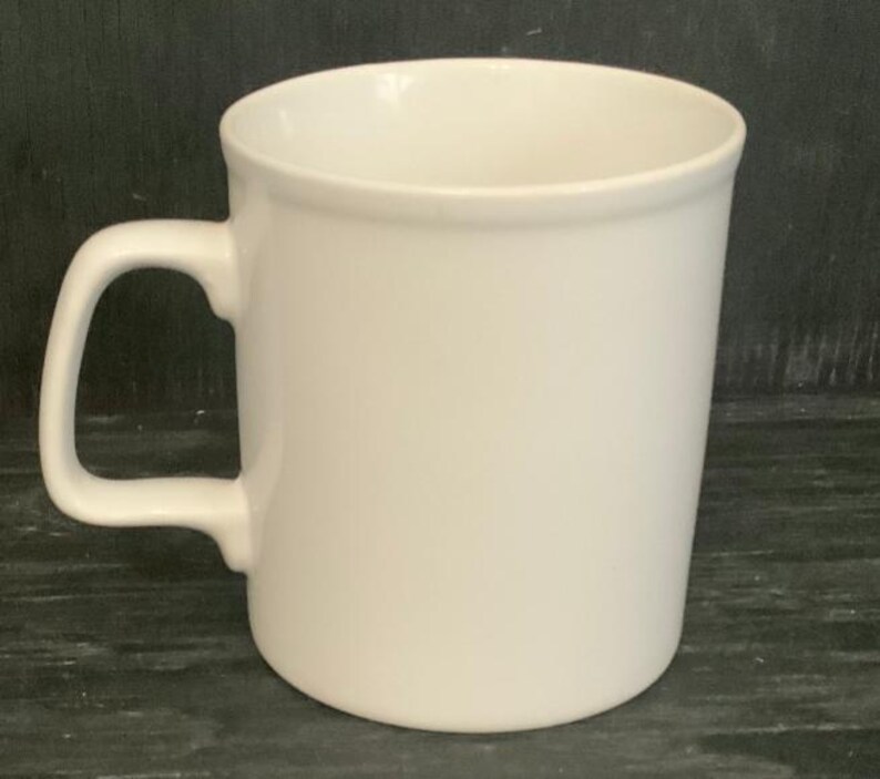 May include: A plain white ceramic mug with a handle. The mug is empty and sitting on a dark surface.