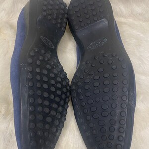 Puede incluir: Un par de mocasines de ante azul con suelas de goma negra. Las suelas tienen un dise&ntilde;o texturizado con peque&ntilde;os bultos redondos para la tracci&oacute;n. Los zapatos se muestran sobre una superficie blanca y esponjosa.