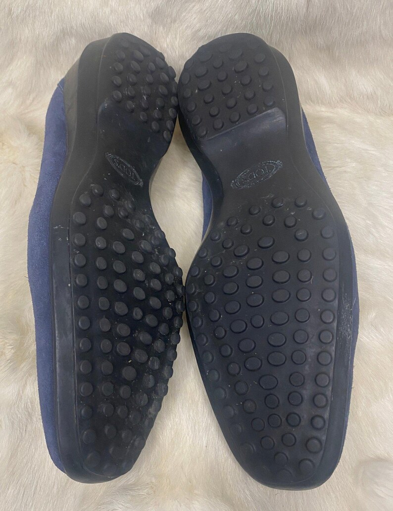 Puede incluir: Un par de mocasines de ante azul con suelas de goma negra. Las suelas tienen un dise&ntilde;o texturizado con peque&ntilde;os bultos redondos para la tracci&oacute;n. Los zapatos se muestran sobre una superficie blanca y esponjosa.