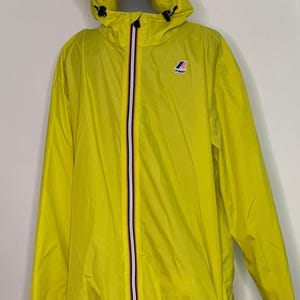 Könnte beinhalten: Hellgelbe Kapuzenjacke mit durchgehendem Reißverschluss und kleinem Logo auf der Brust. Die Jacke hat eine Kapuze mit Kordelzug und ein weißes, rotes und marineblaues Streifendetail am Reißverschluss. Leichte, wasserabweisende Oberbekleidung.