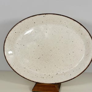 May include: Oval ceramic serving platter with a speckled off-white surface and a dark brown rim. The platter is displayed on a wooden stand. This dish is suitable for serving appetizers or entrees.