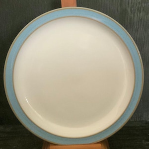 Puede incluir: Un plato de cena de cerámica blanca con un borde azul claro. El plato es redondo y tiene una superficie lisa.