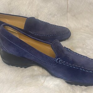 Puede incluir: Un par de mocasines de ante azul marino con tac&oacute;n de cu&ntilde;a bajo. Los zapatos tienen un dise&ntilde;o cl&aacute;sico sin cordones con costuras decorativas y suela negra. Los zapatos se exhiben sobre una superficie blanca texturizada.