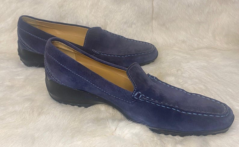 Puede incluir: Un par de mocasines de ante azul marino con tac&oacute;n de cu&ntilde;a bajo. Los zapatos tienen un dise&ntilde;o cl&aacute;sico sin cordones con costuras decorativas y suela negra. Los zapatos se exhiben sobre una superficie blanca texturizada.