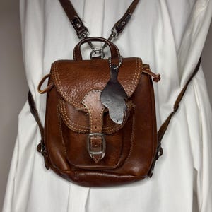 May include: A brown leather backpack featuring a front pocket with a silver-toned metal buckle closure, a top handle, and adjustable shoulder straps. A small, dark, leaf-shaped leather tag hangs from a silver ball chain attached to the top flap.