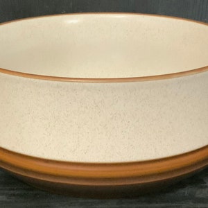 Puede incluir: Un cuenco de cerámica con interior color crema y exterior moteado. El cuenco tiene una banda marrón alrededor de la base y un borde marrón. Es un artículo de menaje.