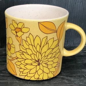 May include: A yellow ceramic mug with a floral pattern in shades of yellow and brown. The mug has a large handle and a speckled glaze.