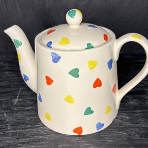 May include: A white ceramic teapot with a lid and a handle. The teapot is decorated with colorful hearts in shades of red, yellow, blue, and green.