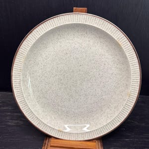 Puede incluir: Un plato redondo de cerámica blanquecina con una superficie moteada y un borde marrón. El plato tiene un borde texturizado y acanalado. El plato se exhibe sobre un soporte de madera sobre un fondo oscuro. Este plato es adecuado para servir alimentos.