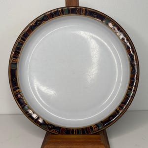 May include: A round, white ceramic plate with a decorative border. The border features a repeating pattern of dark blue, green, and brown shapes. The plate is displayed on a wooden stand.