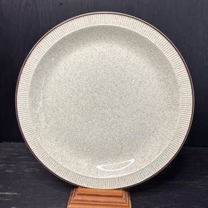 May include: A round, speckled, off-white ceramic plate with a brown rim and a textured, ribbed border. The plate is set on a small wooden stand. This vintage dish is ideal for serving meals. The plate's diameter is approximately 25 cm.