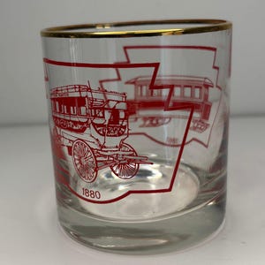May include: Clear glass tumbler with a gold rim and red illustrations of a horse-drawn carriage and a train car. The year "1880" is printed in red on the glass. The glass is cylindrical and sits on a white surface.