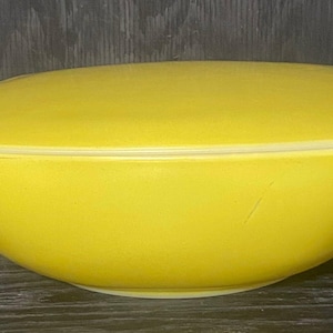 Puede incluir: Un cuenco amarillo brillante con tapa a juego. El cuenco tiene forma redondeada con un borde ligeramente ensanchado. La tapa encaja perfectamente, con pequeñas pestañas para facilitar su extracción. El cuenco es de material liso y sólido.