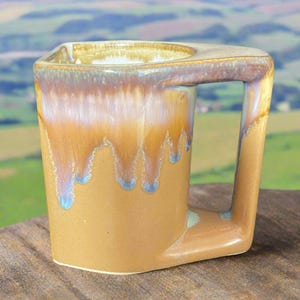 May include: A handmade ceramic mug with an angular design and a built-in handle. The mug has a warm, earthy colour palette with a gradient of brown, orange, and blue hues. The glaze has a dripped effect, adding visual interest to the piece.
