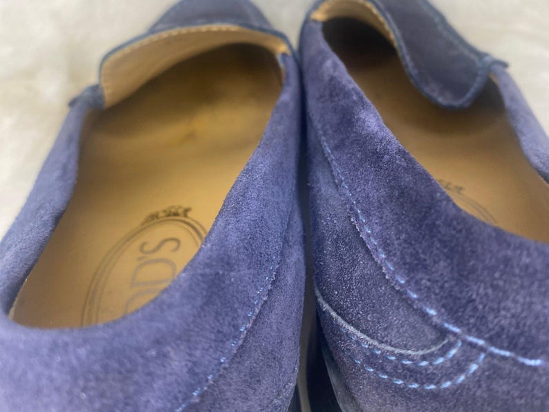 Puede incluir: Primer plano de dos mocasines de ante azul marino. El interior de los zapatos es de color beige claro. La marca "TOD'S" es visible en la plantilla. Los zapatos tienen un dise&ntilde;o cl&aacute;sico sin cordones.