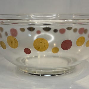 May include: A clear glass mixing bowl featuring a repeating pattern of circular dots in alternating shades of metallic gold and muted red. The bowl has a wide, open top with a smooth, rounded rim and a circular base, showcasing a simple design.