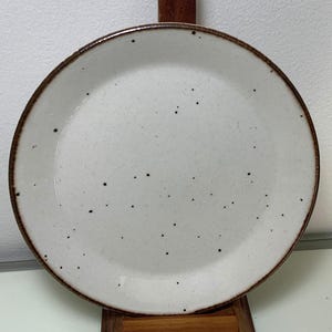 Puede incluir: Un plato de cerámica blanca moteada con un borde marrón. El plato tiene una superficie ligeramente texturizada y se exhibe sobre un soporte de madera. El plato es redondo y tiene un diseño simple y minimalista.