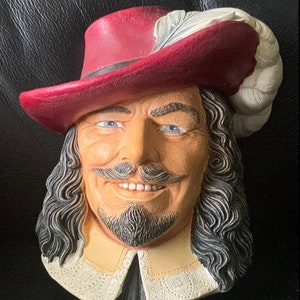 Peut inclure: Une plaque murale en céramique représentant la tête d'un homme portant un chapeau rouge avec une plume blanche. L'homme a une longue perruque noire, une moustache et une barbiche. Il sourit.