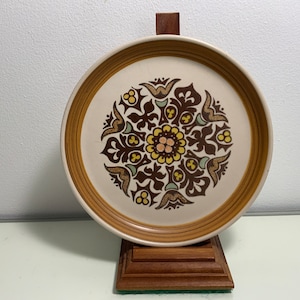 Può includere: Un piatto in ceramica marrone e bianco con un motivo floreale marrone, giallo e verde. Il piatto è su un supporto in legno.