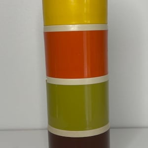 Könnte beinhalten: Ein zylindrischer Vintage-Behälter mit cremefarbenem Deckel und Boden. Der Körper ist in Abschnitte aus Gelb, Orange, Olivgrün und Braun unterteilt, die durch cremefarbene Bänder getrennt sind. Der Behälter ist wahrscheinlich aus Kunststoff.