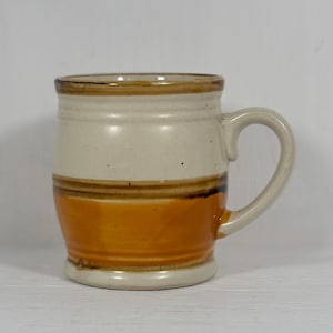 May include: A ceramic mug featuring a two-tone design with a cream-coloured upper section and a warm orange lower section. The mug has a rounded body, a sturdy handle on the right side, and a dark brown horizontal band separating the two colours.