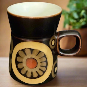 Puede incluir: Una taza de cerámica marrón con un diseño floral amarillo, naranja y verde. La taza tiene un borde ancho y un asa.