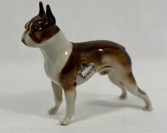 Puede incluir: Figura de cerámica de un perro Boxer, con pecho y cara blancos, y marcas marrones en la cabeza y el cuerpo. El perro está de pie, con la cabeza ligeramente girada hacia la izquierda. Una pequeña etiqueta está adherida a un lado.